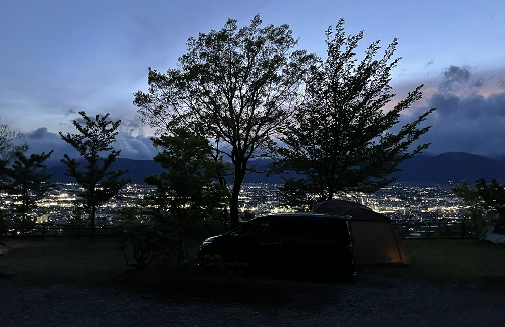 高台のオートキャンプサイトから街の夜景を眺める様子